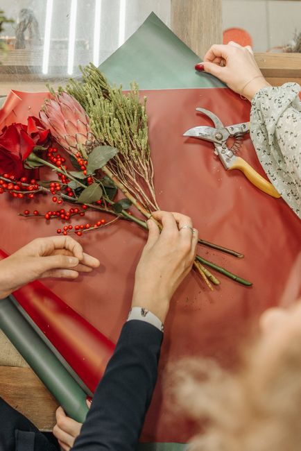 Close-up of a floral arrangement process showing a person with a navy blazer holding a stem of red berries and eucalyptus leaves, while another person with a floral-patterned sleeve arranges a bunch o