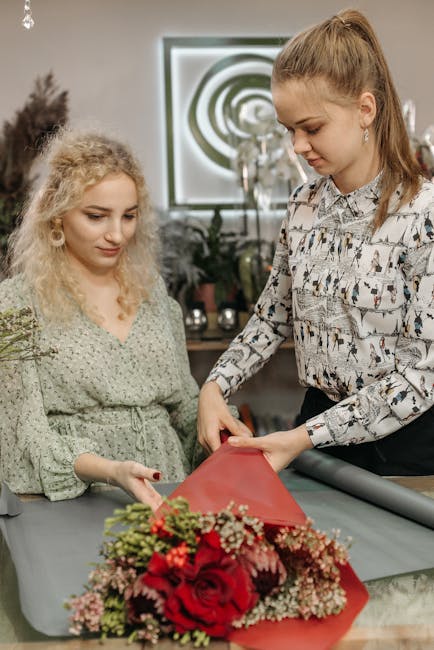 A florist, dressed in a patterned blouse, carefully wraps a vibrant bouquet of red roses, pink baby's breath, and green foliage in red floral wrapping paper. The arrangement is placed on a light grey 