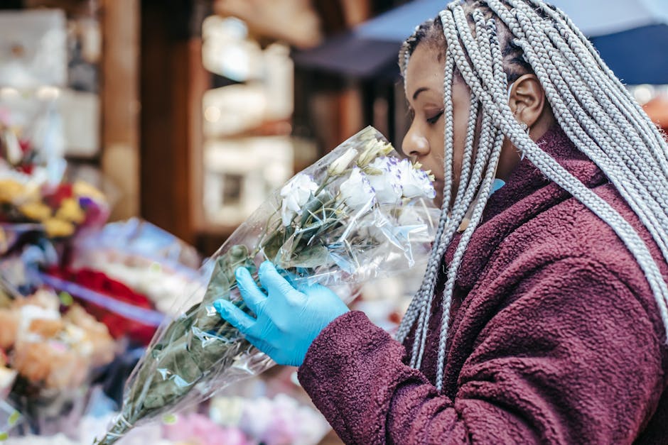 A woman with long, braided silver hair is holding a bouquet of white roses and eucalyptus leaves wrapped in clear plastic. She is wearing a maroon fleece jacket and blue disposable gloves, smelling th