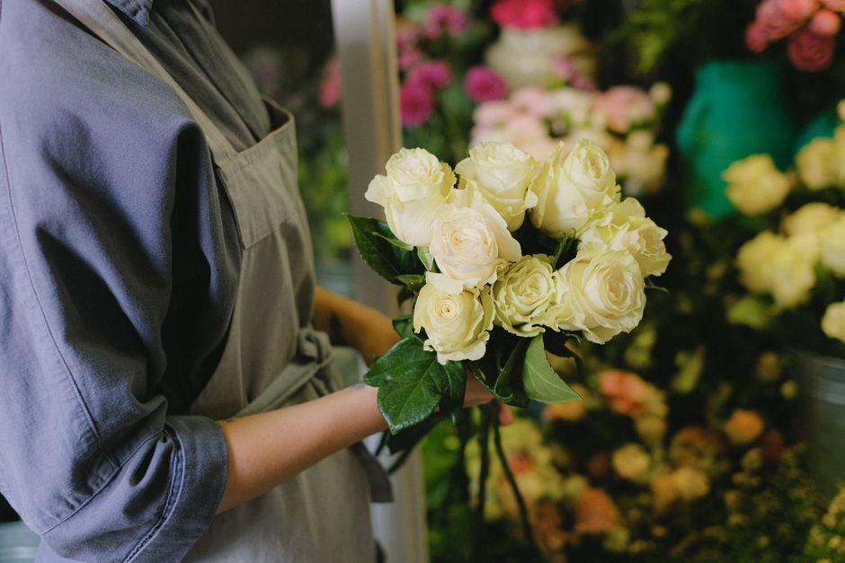 A person wearing a gray apron holds a bouquet of fresh, creamy white roses with dark green leaves, arranged in a rounded bouquet style. The roses are in full bloom with velvety petals, presenting a de